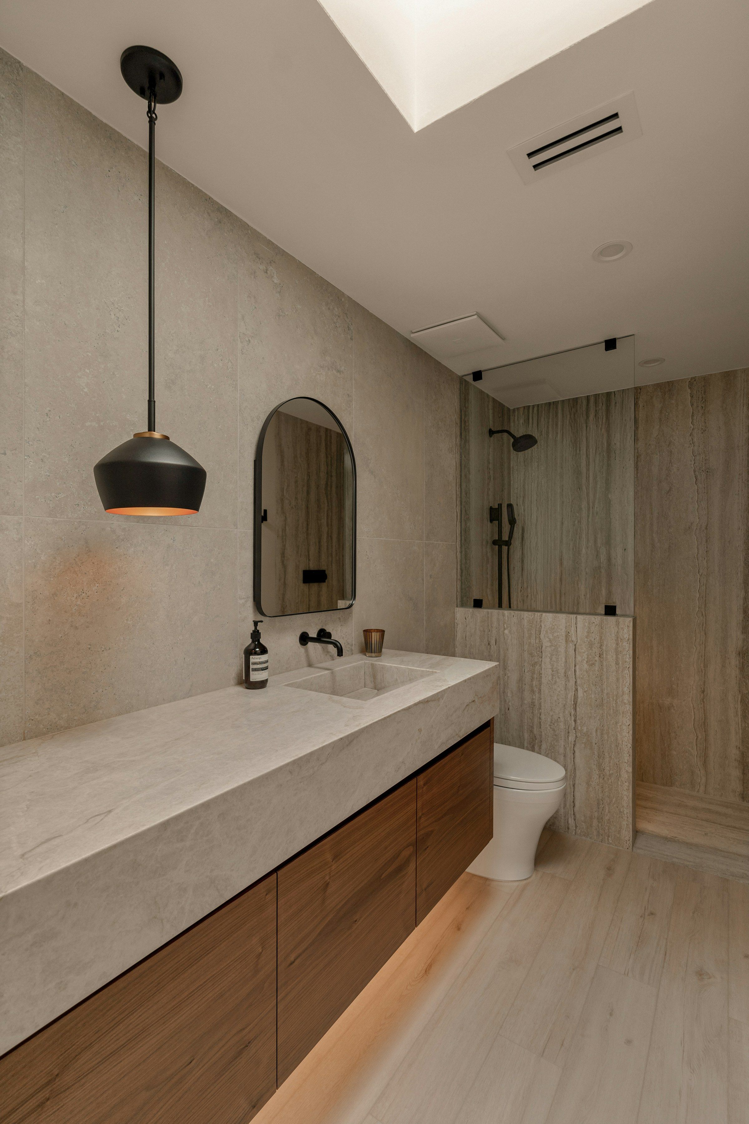 Modernes Bad in hellem Naturstein mit Waschtisch, Holzunterschrank, Pendelleuchte und offener Regendusche