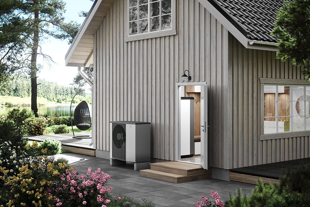 Wärmepumpe vor einem Holzhaus am See, durch die offene Tür ist das Innengerät zu sehen